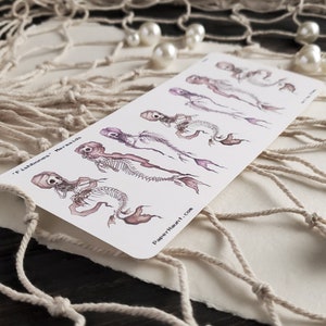 May include: A sheet of temporary tattoos featuring five skeletal mermaids with purple and pink hues. The tattoos are arranged in a row from smallest to largest. The text 'Fishbones' Mermaids' is printed at the top of the sheet. The text 'PaperHaunt.com' is printed at the bottom of the sheet.