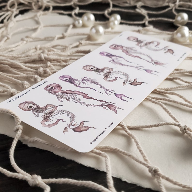 May include: A sheet of temporary tattoos featuring five skeletal mermaids with purple and pink hues. The tattoos are arranged in a row from smallest to largest. The text 'Fishbones' Mermaids' is printed at the top of the sheet. The text 'PaperHaunt.com' is printed at the bottom of the sheet.