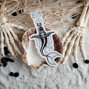 Spooky cute goth Jelly bones mermaid skeleton emerging from water, a sticker Featuring a Mermaid with Translucent skin with visible skeleton and organs resting on a sea shell with skeleton hands.