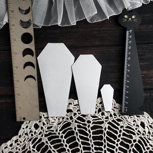 May include: Three white coffin-shaped paper cutouts of varying sizes are displayed on a dark wooden surface, alongside a wooden ruler with moon phase cutouts and a black cat-shaped ruler. A cream-colored lace doily and white tulle fabric are also visible.