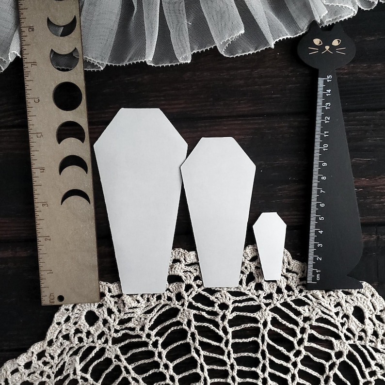 May include: Three white coffin-shaped paper cutouts of varying sizes are displayed on a dark wooden surface, alongside a wooden ruler with moon phase cutouts and a black cat-shaped ruler. A cream-colored lace doily and white tulle fabric are also visible.