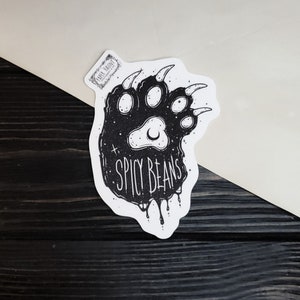 Puede incluir: Una pegatina en blanco y negro con la huella de una pata de oso y el texto "SPICY BEANS". La huella de la pata tiene un dise&ntilde;o de luna creciente y estrellas. La pegatina est&aacute; sobre una superficie de madera blanca y negra.
