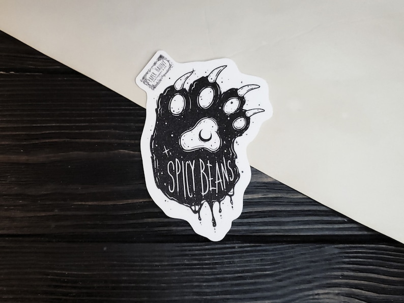 Puede incluir: Una pegatina en blanco y negro con la huella de una pata de oso y el texto "SPICY BEANS". La huella de la pata tiene un dise&ntilde;o de luna creciente y estrellas. La pegatina est&aacute; sobre una superficie de madera blanca y negra.