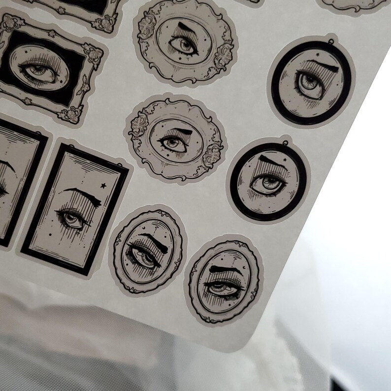 May include: A sheet of stickers featuring black and white illustrations of eyes in various ornate frames. The stickers are on a white background.