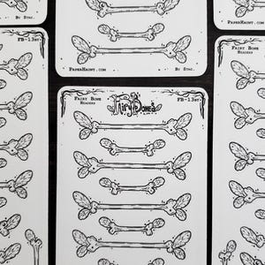 May include: A collection of white stickers featuring black illustrations of fairy bones. The stickers have a bone-like shape with leaf-like details at the ends. The text "Fairy Bones" and "FB-13BT" is visible on the stickers. The website "PAPERHAUNT.COM" is also visible.
