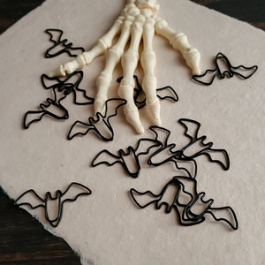 May include: A white plastic skeleton hand with black metal bat-shaped paper clips scattered around it on a beige background.
