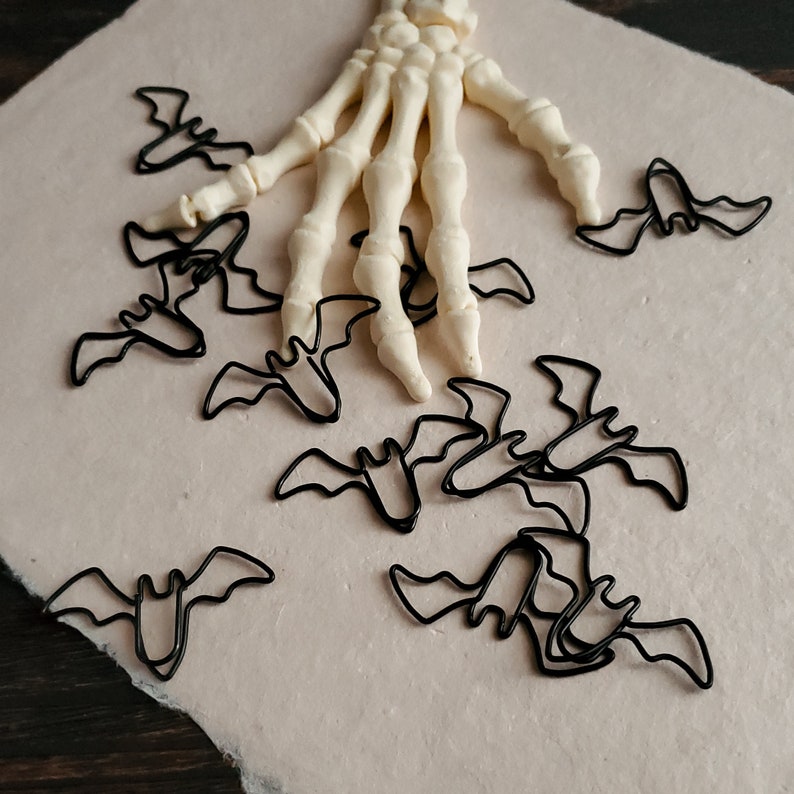May include: A white plastic skeleton hand with black metal bat-shaped paper clips scattered around it on a beige background.