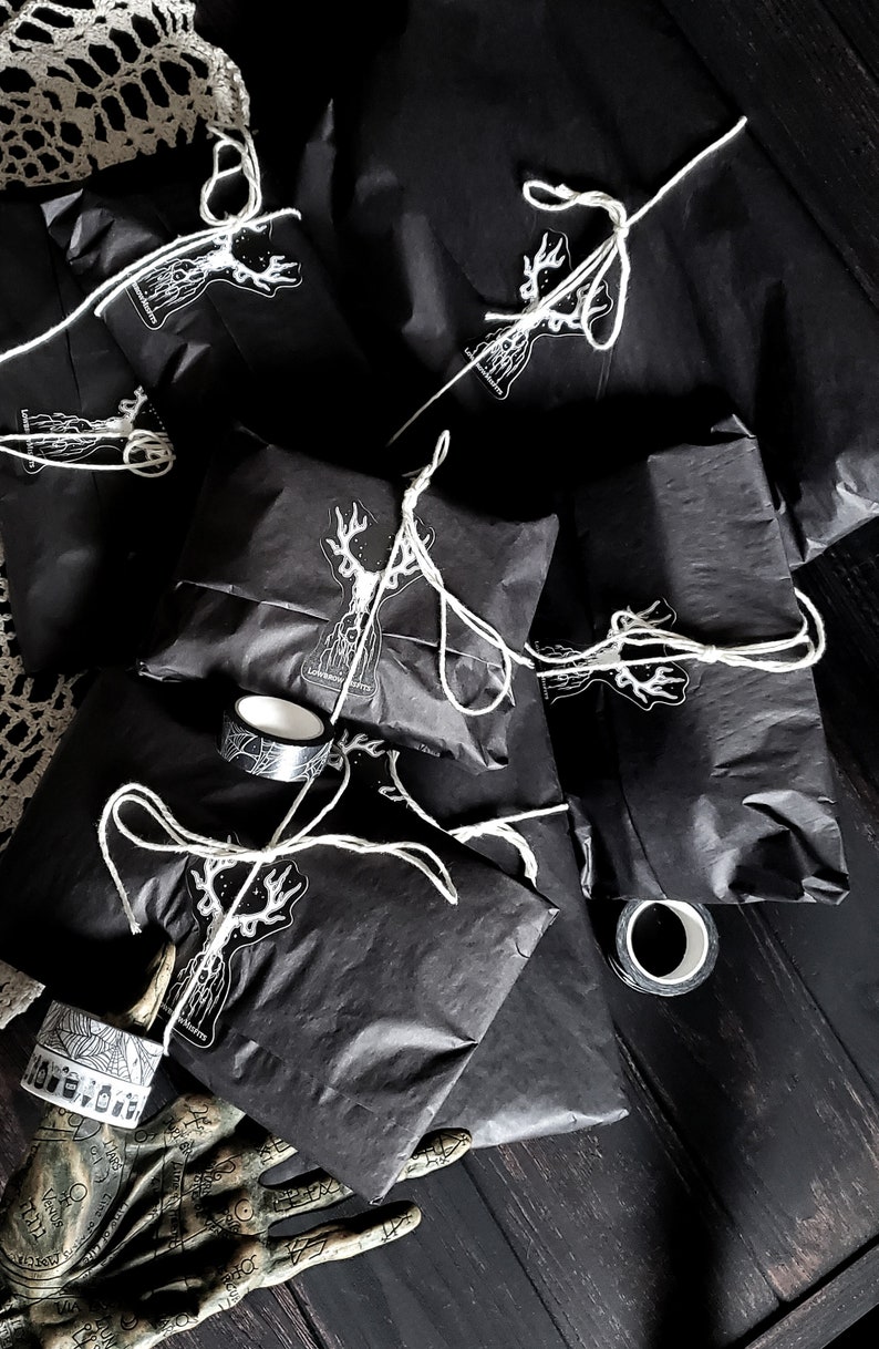 May include: A pile of black gift bags with white string and a white deer sticker on each bag. The bags are on a dark wooden surface with a decorative hand and a roll of washi tape.