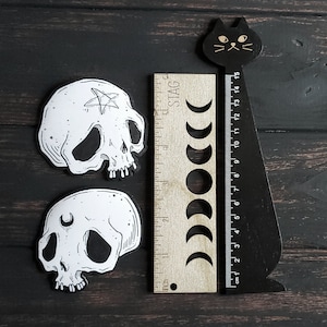 Op de afbeelding: Twee zwart-witte schedelstickers met een pentagram en een halve maan ontwerp. De stickers zijn op een donker houten achtergrond met een zwarte kat liniaal.