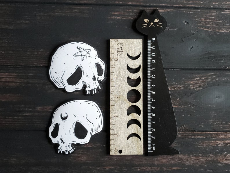 Op de afbeelding: Twee zwart-witte schedelstickers met een pentagram en een halve maan ontwerp. De stickers zijn op een donker houten achtergrond met een zwarte kat liniaal.