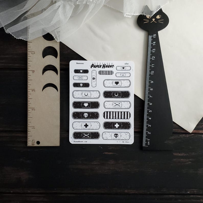 May include: A sheet of black and white patterned bandaid stickers. The stickers feature various designs including hearts, skulls, crosses, and crescent moons. The sheet is labeled "PAPER HAUNT".
