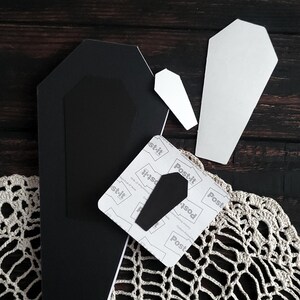 May include: A collection of Halloween-themed craft supplies, including black and white coffin shapes, a Post-it note with a coffin cutout, and a crocheted lace doily. The items are arranged on a dark wooden surface.