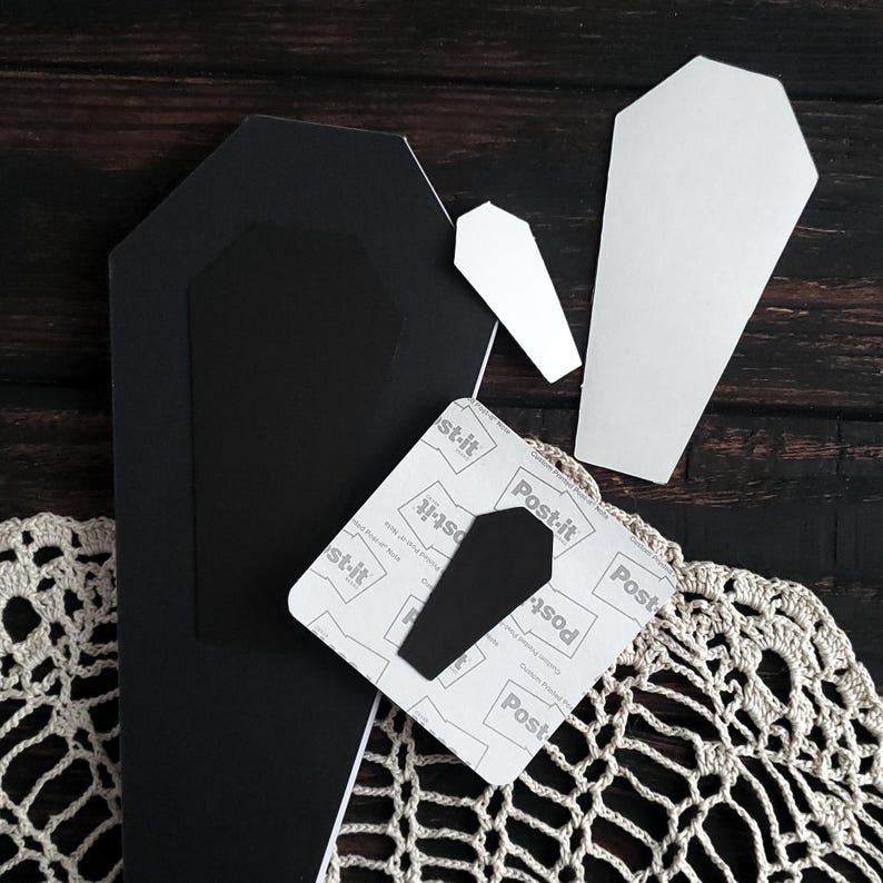 May include: A collection of Halloween-themed craft supplies, including black and white coffin shapes, a Post-it note with a coffin cutout, and a crocheted lace doily. The items are arranged on a dark wooden surface.