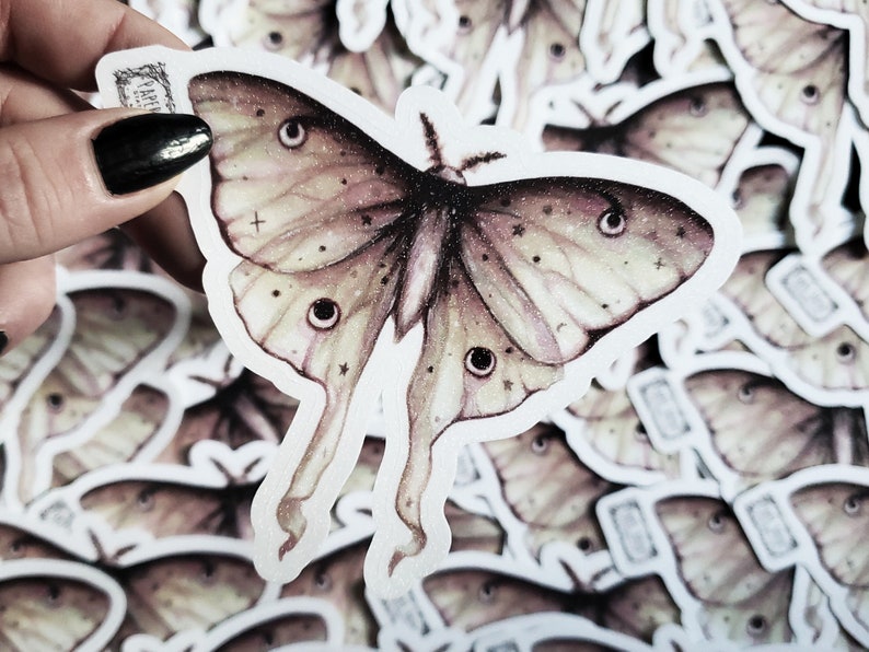 May include: A close-up of a sticker featuring a detailed illustration of a pale green and pink moth with black eyes and a white background. The moth has a delicate, ethereal appearance and is surrounded by other stickers.