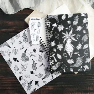 May include: A black and white spiral bound notebook with a black cover featuring a repeating pattern of leaves, mushrooms, and a humanoid figure with a root-like body. The notebook is open to reveal a white page with a similar pattern. The text "PAPER HAUNT" is visible on a sticker.