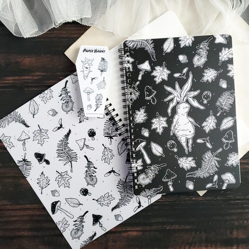 May include: A black and white spiral bound notebook with a black cover featuring a repeating pattern of leaves, mushrooms, and a humanoid figure with a root-like body. The notebook is open to reveal a white page with a similar pattern. The text "PAPER HAUNT" is visible on a sticker.