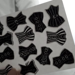May include: A sheet of black and white corset stickers. The corsets are in various styles, some with lace, some with stripes, and some with buckles. The stickers are on a clear background.