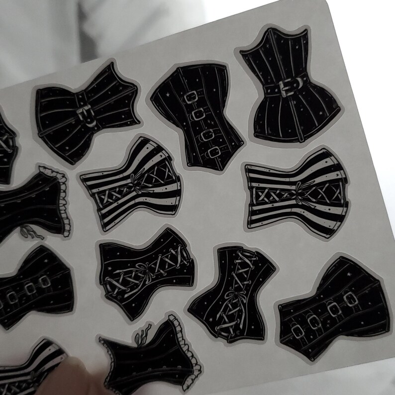 May include: A sheet of black and white corset stickers. The corsets are in various styles, some with lace, some with stripes, and some with buckles. The stickers are on a clear background.