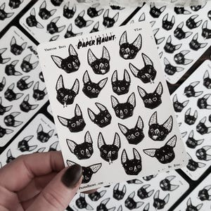 May include: A sheet of stickers featuring black and white illustrations of bats with various expressions. The stickers are arranged in a grid pattern and have a white background. The sheet is labeled "Vampire Bats" and "Paper Haunt."