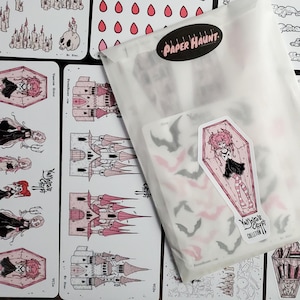 May include: A clear plastic envelope with the text "PAPER HAUNT." printed on it. The envelope contains a set of stickers featuring a pink coffin with a vampire girl inside, bats, and a castle. The stickers are part of a "Vampire's Crypt Collection."