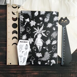 May include: A black and white spiral bound notebook featuring a whimsical illustration of a forest creature with leaves and mushrooms. The notebook comes with a sticker sheet featuring a similar design. The sticker sheet is labelled "Paper Haunt."