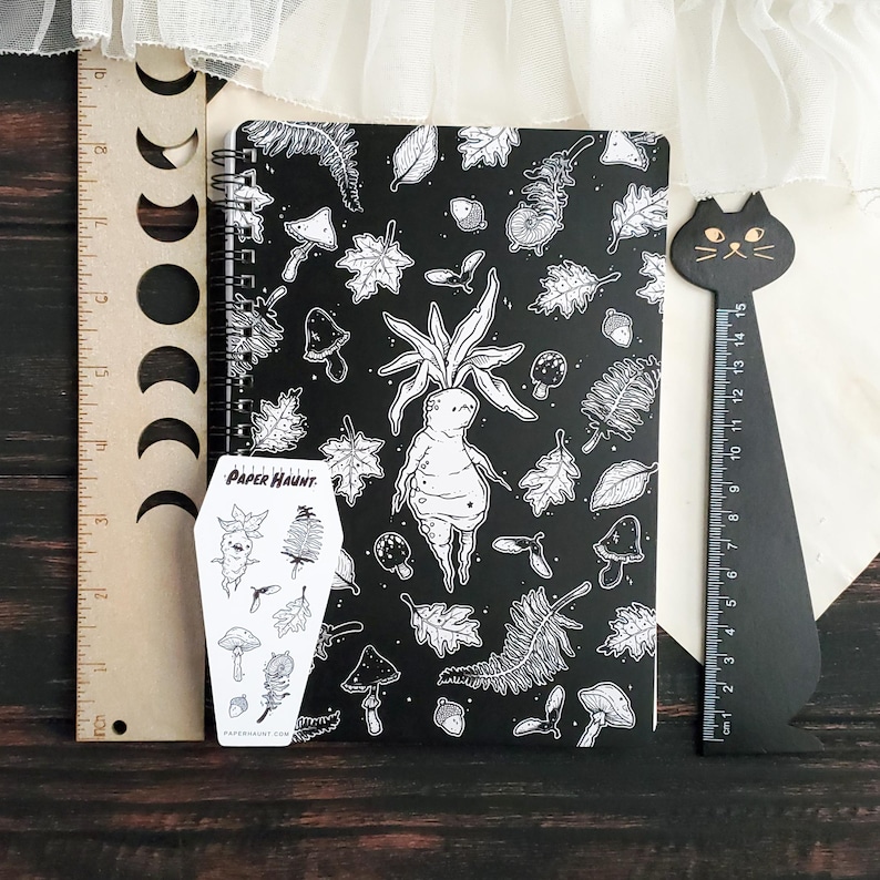 May include: A black and white spiral bound notebook featuring a whimsical illustration of a forest creature with leaves and mushrooms. The notebook comes with a sticker sheet featuring a similar design. The sticker sheet is labelled "Paper Haunt."