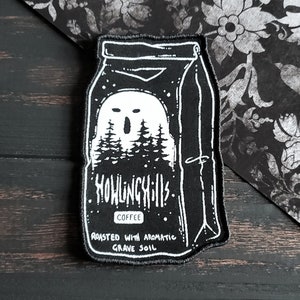 May include: Black and white embroidered patch of a coffee bag with a ghost inside. The bag has the text "Howling Hills" and "Roasted with aromatic grave soil" on it.