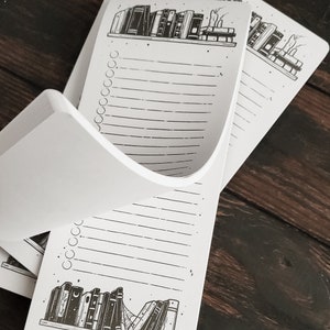 K&ouml;nnte beinhalten: Ein wei&szlig;er Notizblock mit einer Schwarzwei&szlig;illustration eines B&uuml;cherregals voller B&uuml;cher. Der Notizblock hat einen linierten Bereich zum Schreiben und einen Bereich mit Kreisen zum Abhaken von Aufgaben.