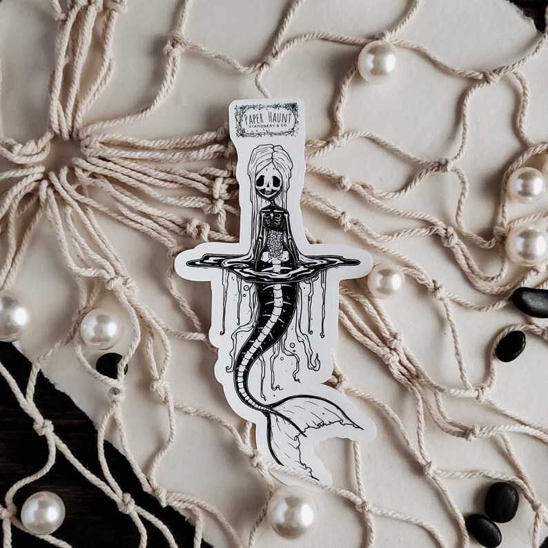 Spooky cute goth Jelly bones mermaid skeleton emerging from water, a sticker Featuring a Mermaid with Translucent skin with visible skeleton and organs resting on a fishing net with black stones and pearls.