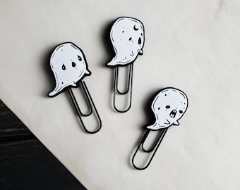May include: Three black and white ghost-shaped paper clips. Each clip has a different ghost face with eyes, a mouth, and other details.