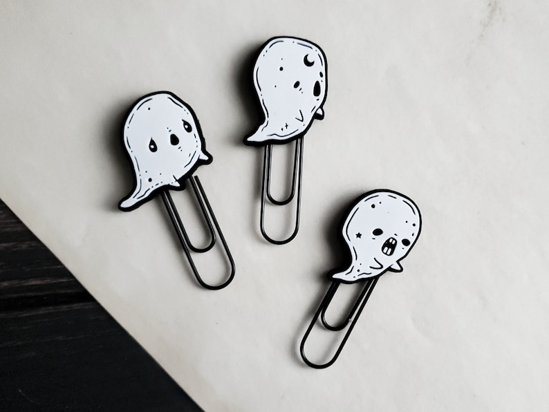 May include: Three black and white ghost-shaped paper clips. Each clip has a different ghost face with eyes, a mouth, and other details.