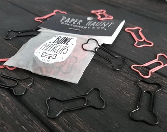 May include: A close-up of a bag of bone-shaped paperclips. The bag is white and has a black label that says "Bone Paperclips" and "Paper Haunt Stationery & Co.". There are also pink bone-shaped paperclips scattered around the bag.