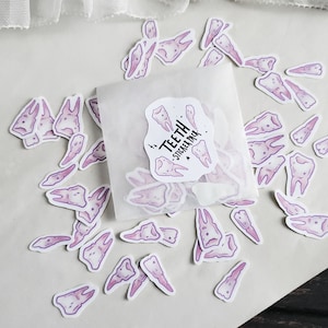 May include: A sheet of white paper with a variety of purple tooth stickers scattered on it. The stickers are in a variety of shapes and sizes. The text "Teeth Sticker Pack" is visible on a sticker in the centre of the image.