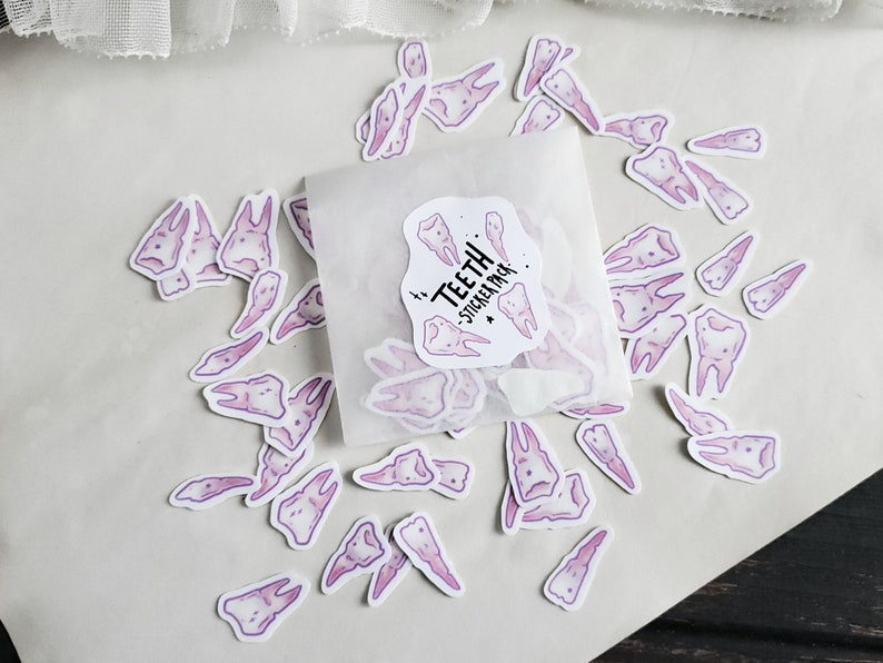 May include: A sheet of white paper with a variety of purple tooth stickers scattered on it. The stickers are in a variety of shapes and sizes. The text "Teeth Sticker Pack" is visible on a sticker in the centre of the image.
