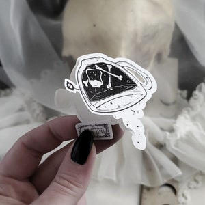 Op de afbeelding: Een zwart-witte sticker van een theepot met een schedel en gekruiste beenderen erin. De theepot giet een stroom stippen uit.