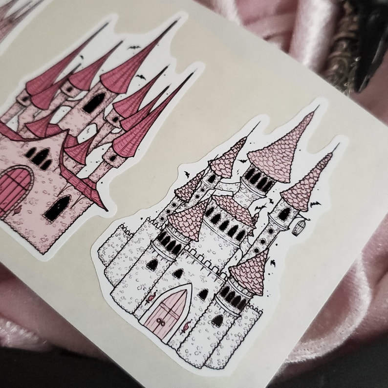 May include: Two pink and white castle stickers with black outlines. The castles have multiple towers and windows. The stickers are on a white background.