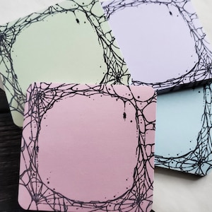 May include: Four square sticky notes with a black spiderweb design border. The notes are in pastel colors: light green, pink, light blue, and lavender.