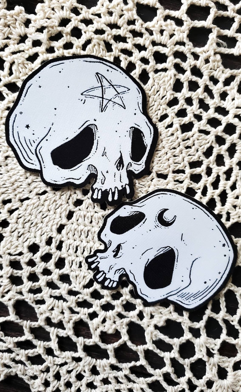 Op de afbeelding: Twee zwart-witte schedel-illustraties met een pentagram en een halve maan op een wit gehaakt oppervlak.