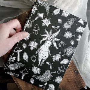 May include: Black and white spiral notebook featuring a whimsical illustration of a forest creature with leaves, mushrooms, and acorns. The notebook has a black cover with a white design.