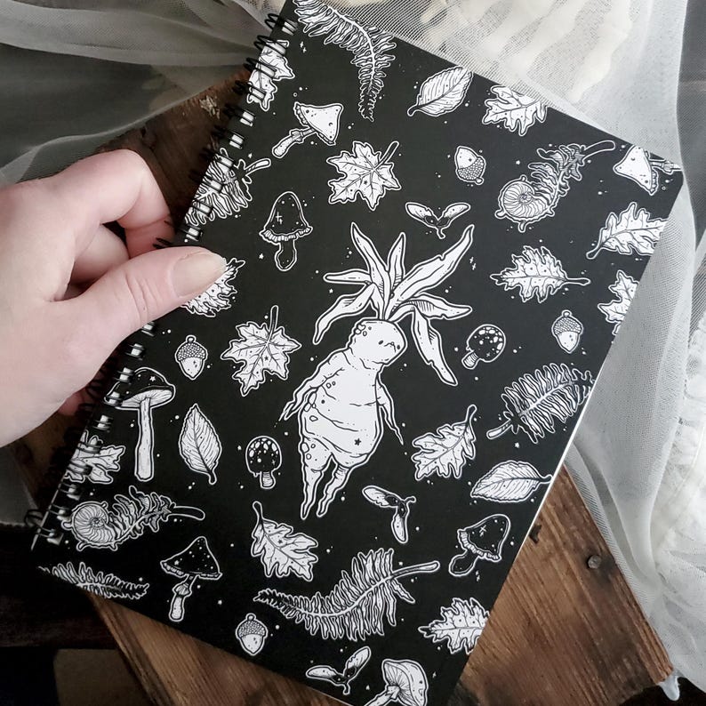 May include: Black and white spiral notebook featuring a whimsical illustration of a forest creature with leaves, mushrooms, and acorns. The notebook has a black cover with a white design.
