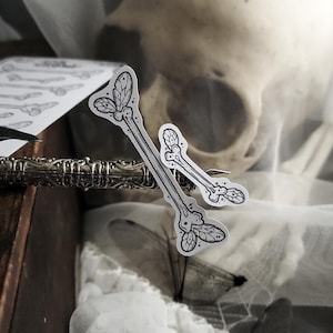 May include: Two bone-shaped stickers with leaf and dot details, in black and white, are displayed. The stickers are on a decorative metal object, with a skull and fabric in the background. The stickers have a gothic or Halloween theme.