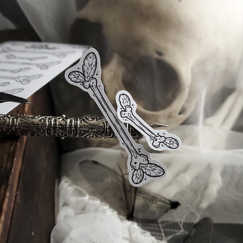 May include: Two bone-shaped stickers with leaf and dot details, in black and white, are displayed. The stickers are on a decorative metal object, with a skull and fabric in the background. The stickers have a gothic or Halloween theme.