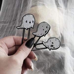 May include: Three black metal ghost-shaped paper clips with white outlines and black eyes. The ghosts are attached to the paper clips with a silver metal ring.