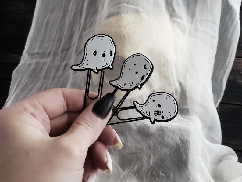 May include: Three black metal ghost-shaped paper clips with white outlines and black eyes. The ghosts are attached to the paper clips with a silver metal ring.