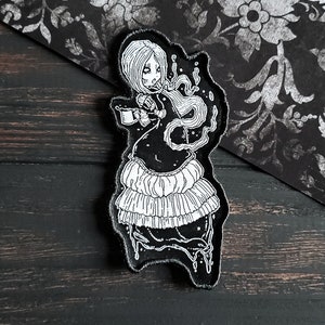May include: Black and white embroidered patch of a woman in a flowing dress holding a cup. The woman has long hair and a crown of flowers. The patch is on a black background.