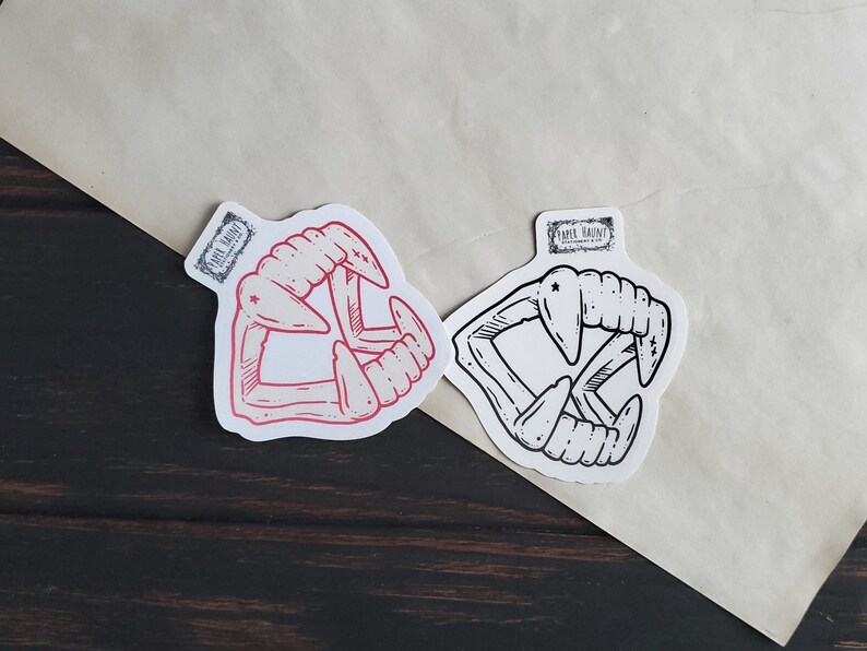 May include: Two sticker designs of a set of fangs, one in red and one in black and white. The stickers are on a white background.
