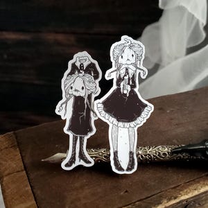 May include: Two black and white sticker illustrations of girls wearing dresses. The girl on the left is holding a doll in her arms. The girl on the right is holding a small object in her hands. Both girls have long hair and are wearing black dresses with white accents.