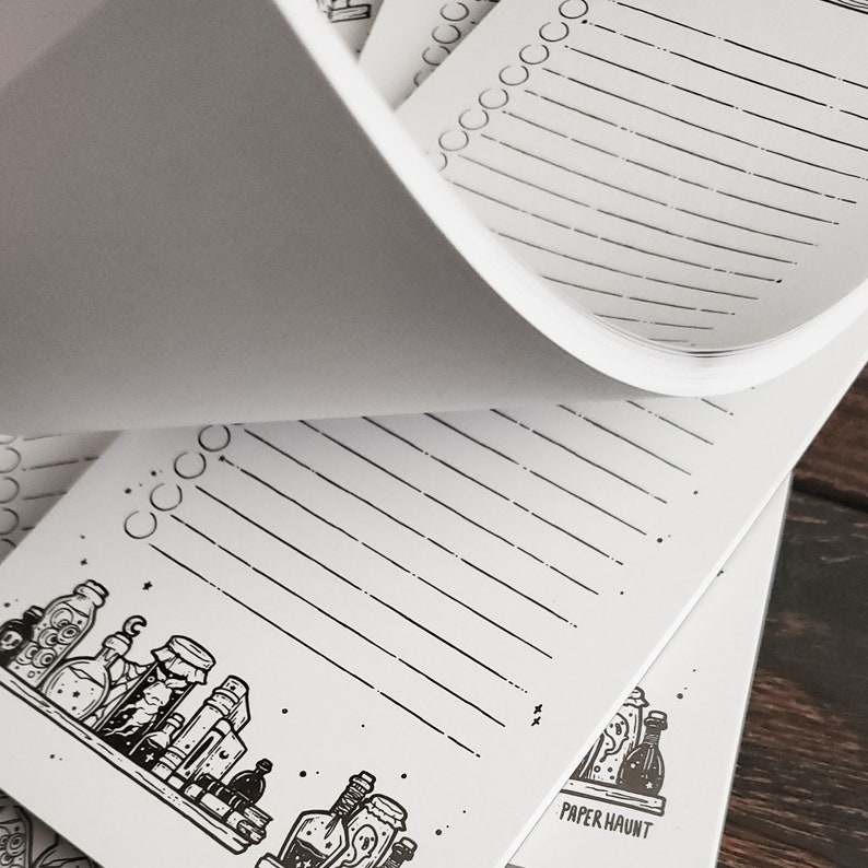 Puede incluir: Un bloc de notas blanco con una ilustraci&oacute;n en blanco y negro de botellas de pociones y el texto "PAPER HAUNT". El bloc de notas tiene p&aacute;ginas rayadas y una lista de verificaci&oacute;n.