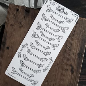 May include: A sheet of stickers featuring bone-shaped designs with leaf-like accents. The stickers are black and white, with the words "Fairy Bone Headers" and "PaperHaunt.com" printed on the sheet. The stickers are arranged on a wooden surface.