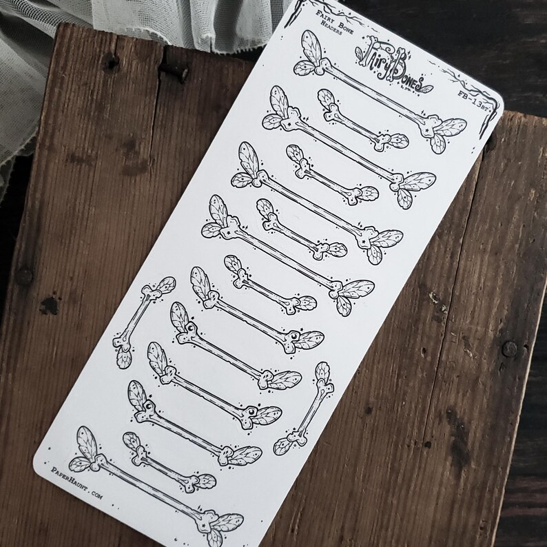 May include: A sheet of stickers featuring bone-shaped designs with leaf-like accents. The stickers are black and white, with the words "Fairy Bone Headers" and "PaperHaunt.com" printed on the sheet. The stickers are arranged on a wooden surface.